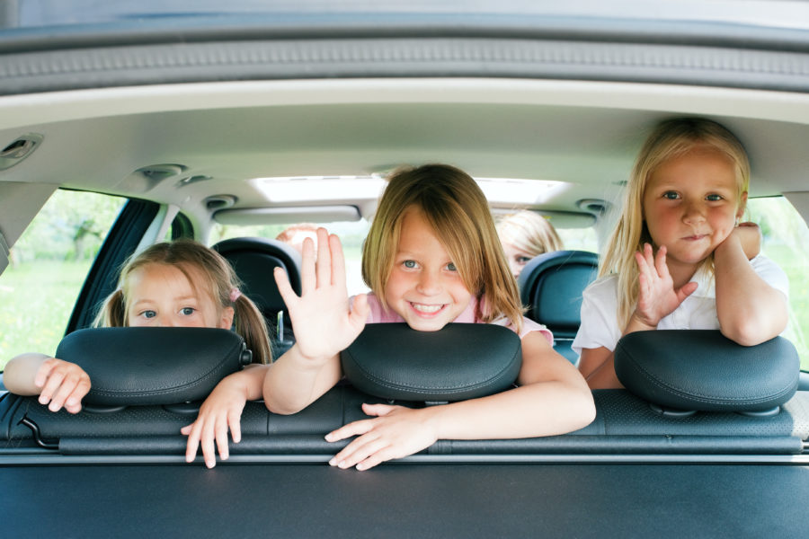 Three little blonde girls facing backwards and waving from the back seat of car