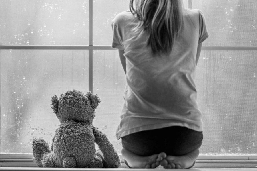 Black and white photo of girl sitting with teddy bear looking out window