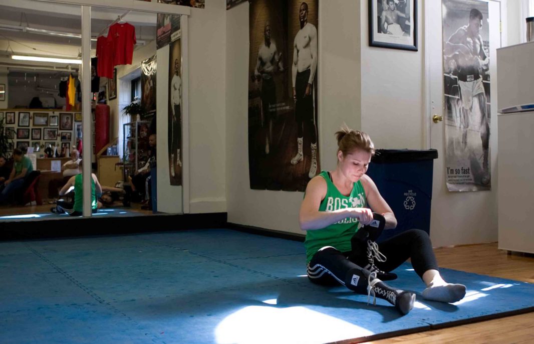 Haymakers of Hope founder Julie Anne Kelly sitting on blue mat and lacing up shoes at the boxing gym