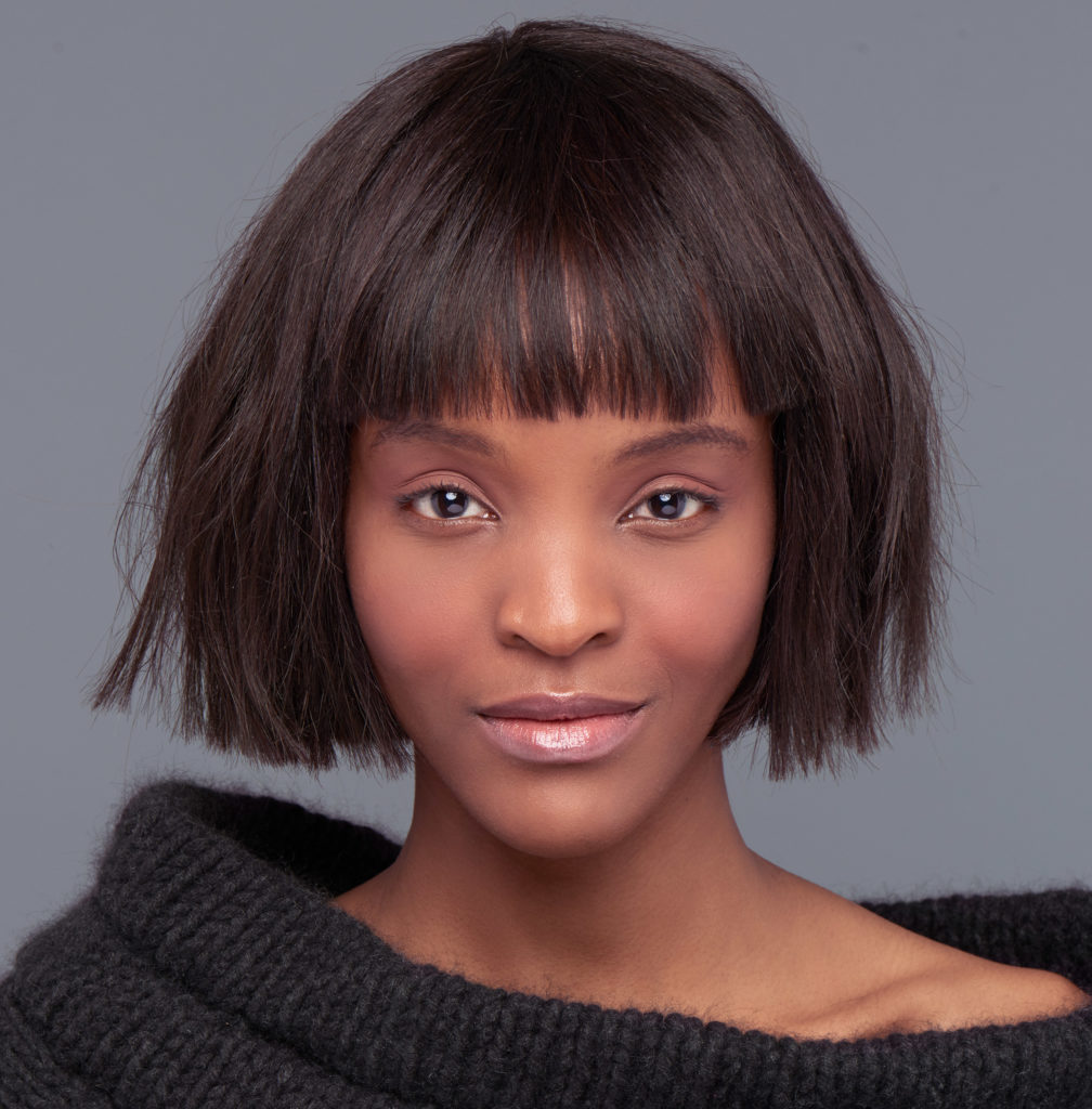 A blunt bob with bangs on model with dark natural hair in a black shoulder-baring sweater 