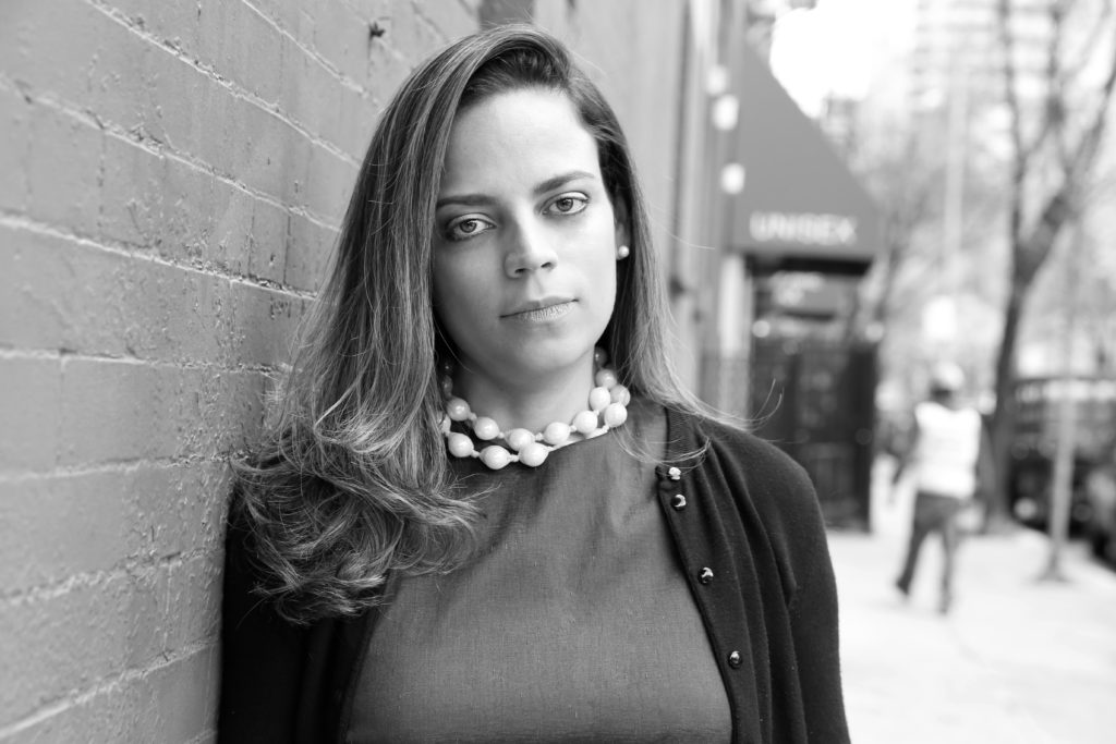Black and white portrait of Fashion designer Melina Cortes-Nmili leaning on brick wall in cardigan and oversize plastic bead necklace