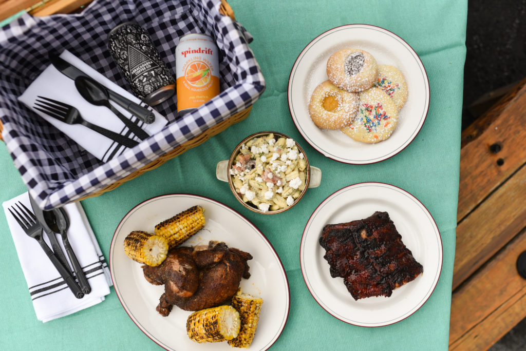picnic basket with gingham interior and plates of chicken, ribs, corn on the cob, cookies on mint-colored cloth