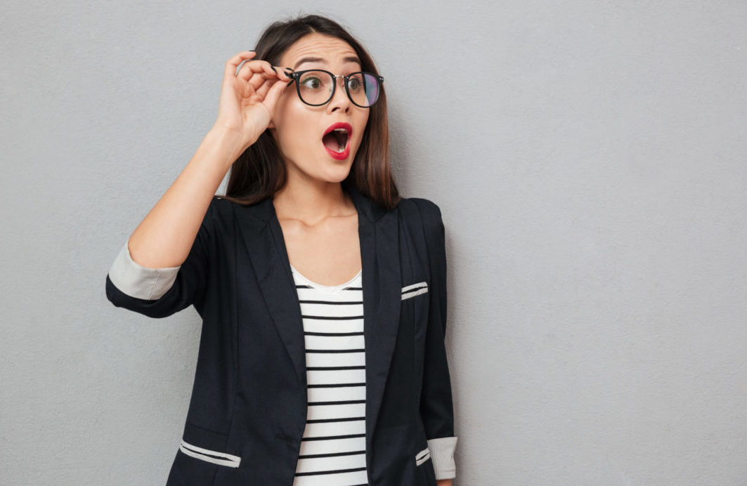 Young woman holding side of eyeglasses and looking surprised in navy blazer and striped shirt