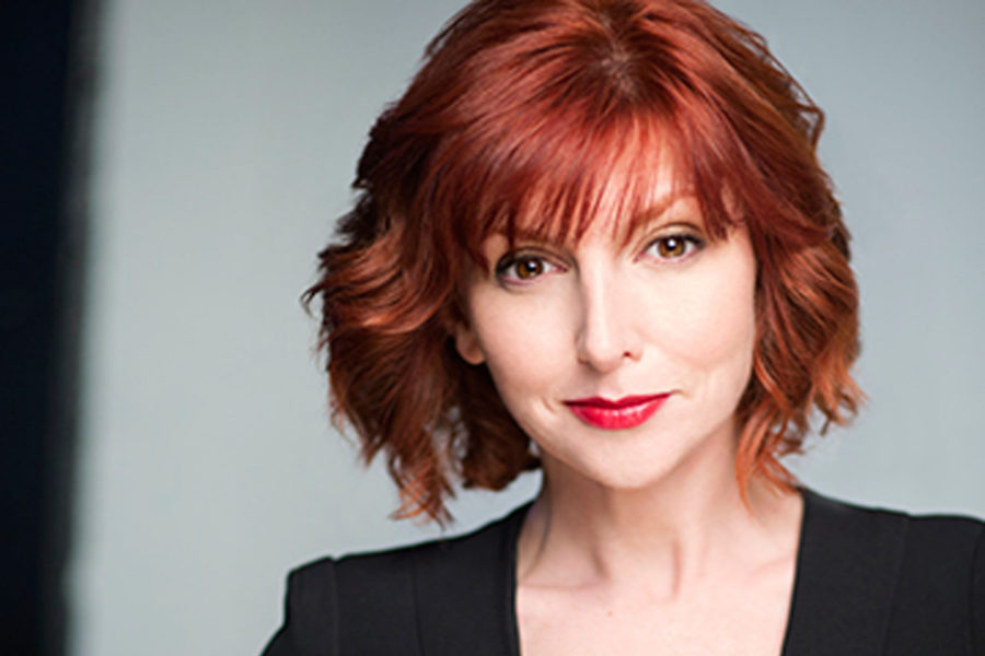 Redheaded comedian Erin Maguire in a black jumpsuit with skinny blue belt