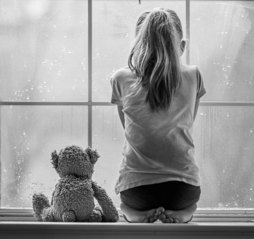 Black and white photo of little girl and teddy bear sitting on window ledge looking outside