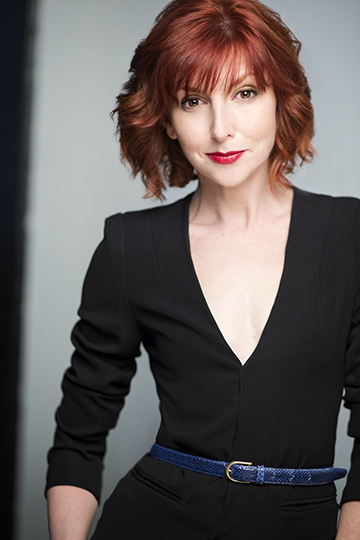 Redheaded comedian Erin Maguire in a black jumpsuit with skinny blue belt