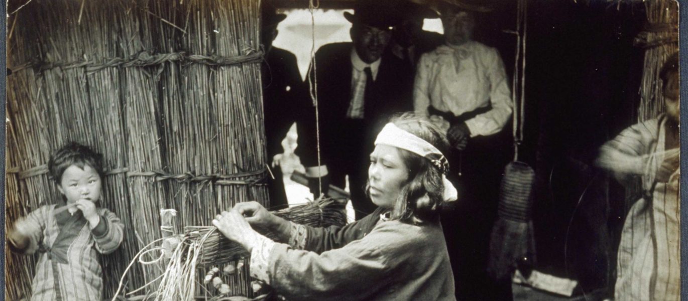 Four “Anglo” fairgoers watching a Japanese woman weave outdoors. woman is seated on a small block of wood; her son stands near her, leaning against the wall of a thatched dwelling, 1904. Photographer: Jessie Tarbox Beals